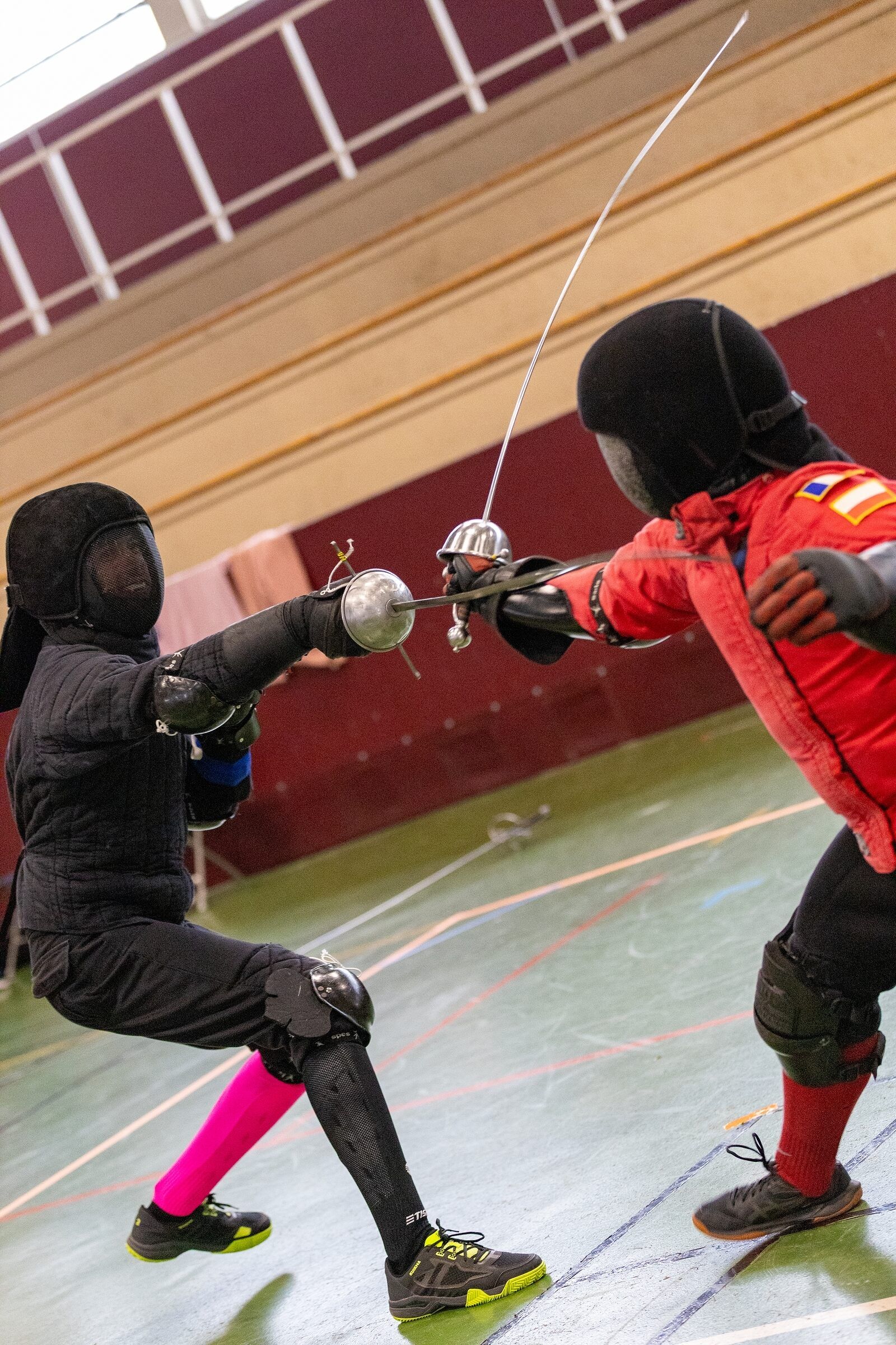 Duel à l'épée de côté entre deux escrimeurs, en noir et en rouge, dans une salle aux murs bordeaux.