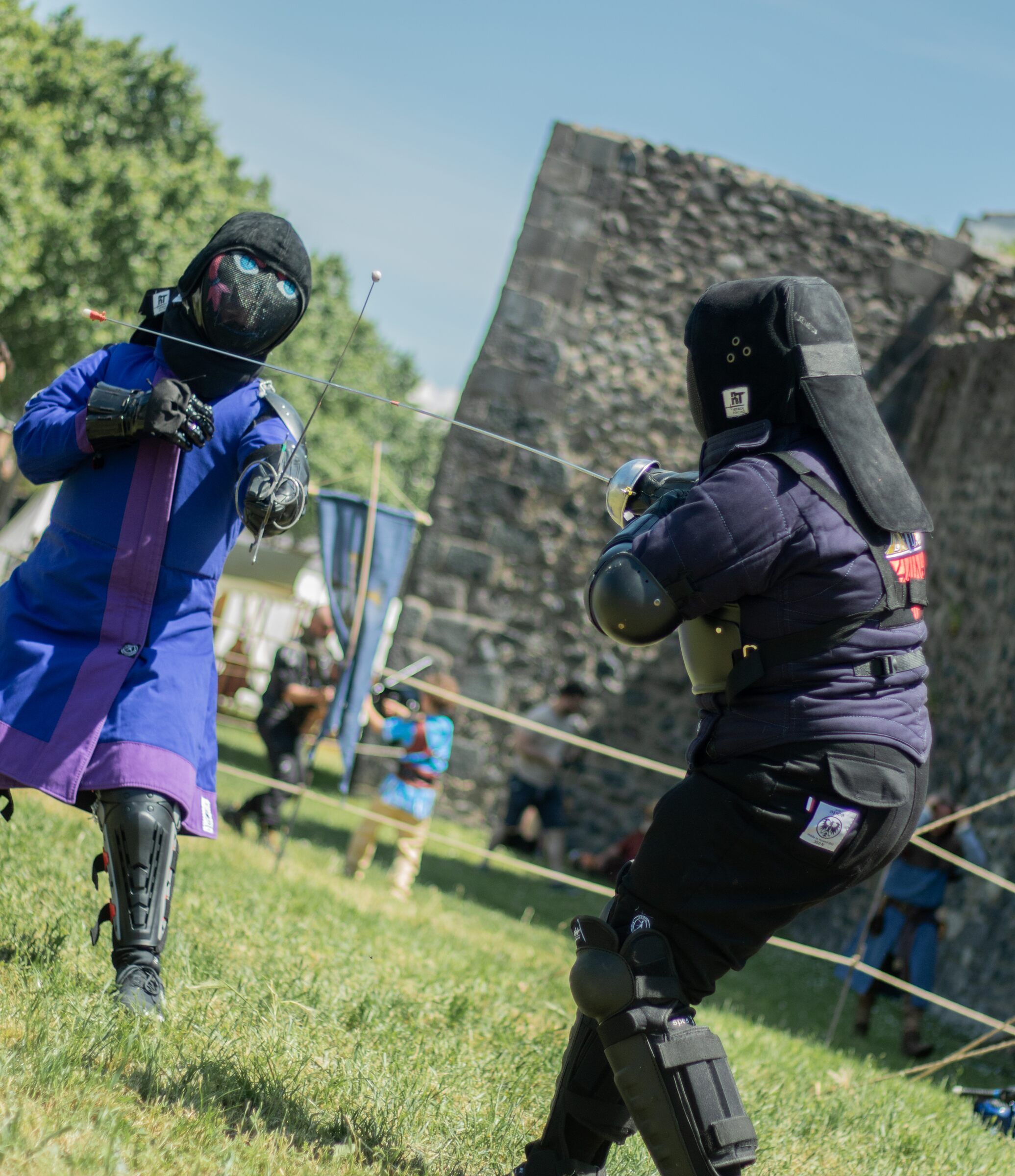 Deux escrimeurs s'affrontent sur l'herbe devant des remparts.