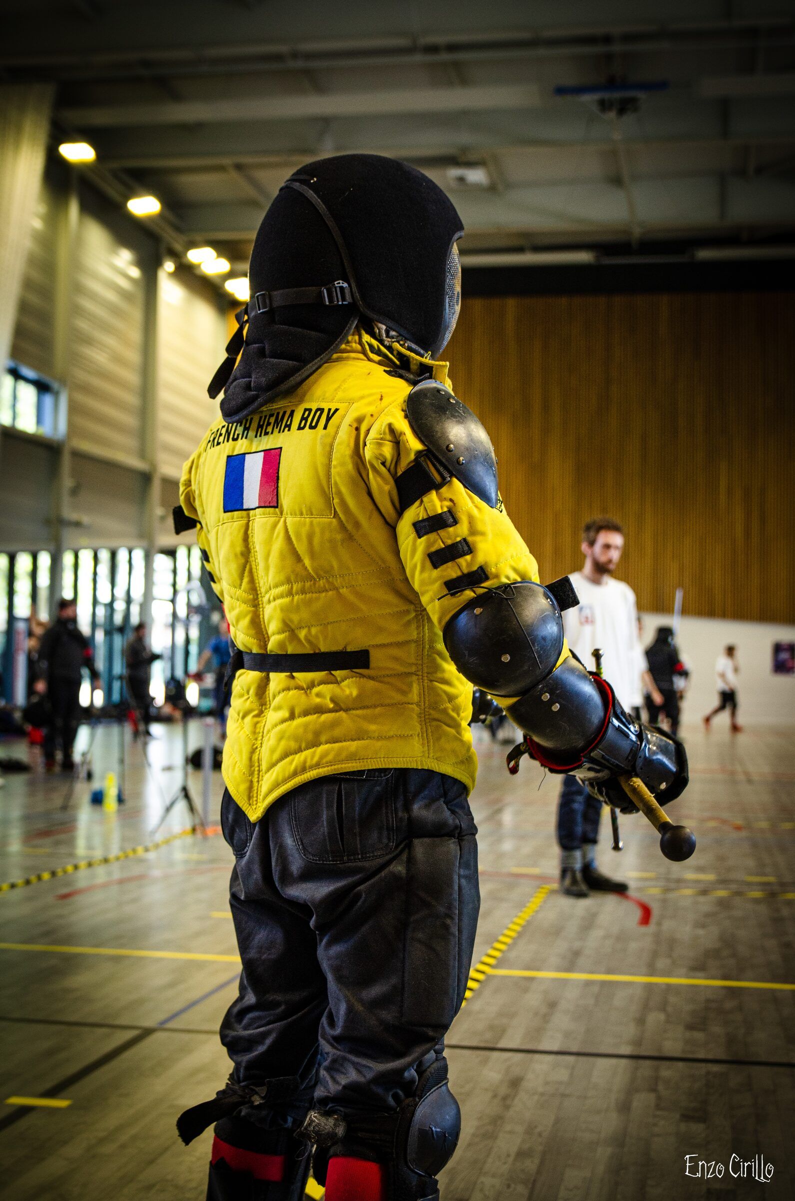 Gabriel Tardio de dos sur l'aire de tournoi, gambison jaune, épée longue tenue basse.
